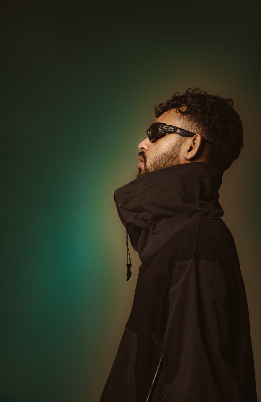 Clover — Suvin Somanna in side profile, wearing sunglasses and a black jacket in green and amber studio light